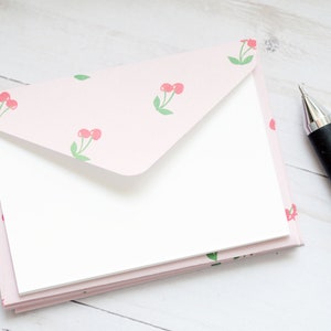 May include: A stack of stationery with a light pink cherry pattern. The stationery includes a matching envelope and blank white cards. A black and silver pen rests on the side, ready for writing.