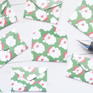 May include: Several small, green envelopes featuring a Santa Claus face pattern. The envelopes are arranged on a white surface, with a pen. Perfect for Christmas or seasonal greetings.