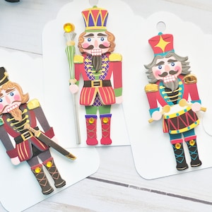 May include: Three colorful nutcracker gift tags with different designs. One nutcracker holds a drum, another holds a staff, and the third holds a sword. The tags are white with a scalloped edge.