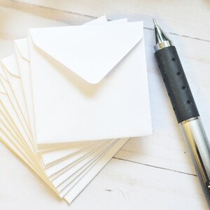 May include: A stack of white envelopes with a silver pen on a white wooden surface. The pen has a black rubber grip and the word "TUL" printed on the side.
