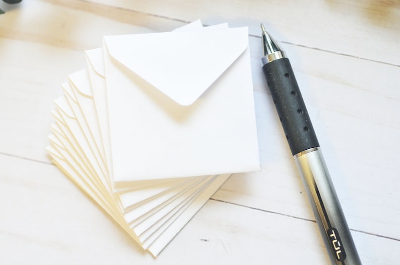 May include: A stack of white envelopes with a silver pen on a white wooden surface. The pen has a black rubber grip and the word "TUL" printed on the side.