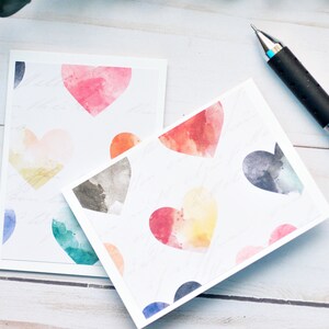 May include: Two white cards with watercolor hearts in various colors. The cards are on a white wooden surface with a pen.