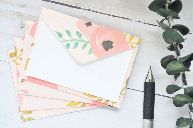 May include: A stack of five pink envelopes with a floral pattern. The envelopes have a white interior and are arranged in a fan shape. The floral pattern includes pink flowers, green leaves, and gold accents.