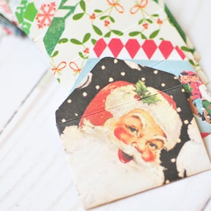 May include: A collection of Christmas-themed envelopes with various festive patterns and illustrations. One envelope features Santa Claus, while others display holiday designs with red, green, and white colors. The envelopes are arranged in a fan shape.