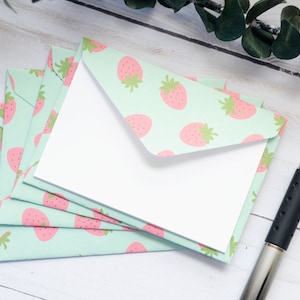 May include: A stack of light blue envelopes with a pink strawberry pattern. The envelopes are blank and ready to be addressed. A silver pen is lying on a white wooden surface.