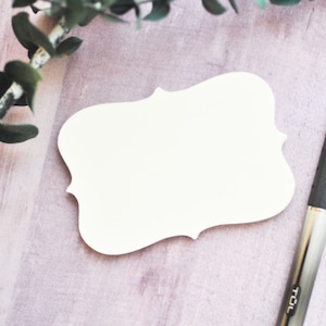 May include: A white, blank, decorative, paper tag with a scalloped edge. The tag is on a light pink wooden surface with a green leafy branch and a silver pen.
