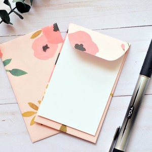 May include: Two pink floral patterned envelopes with a white envelope inside. A silver pen with a black rubber grip is on a white wooden surface.