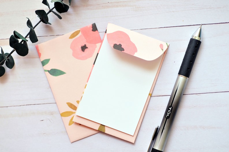 May include: Two pink floral patterned envelopes with a white envelope inside. A silver pen with a black rubber grip is on a white wooden surface.