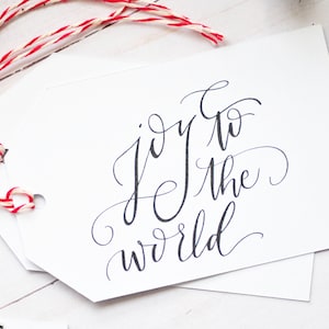 May include: White gift tags with the phrase "joy to the world" in elegant black calligraphy. The tags are strung with red and white twine, perfect for holiday or Christmas gifts.
