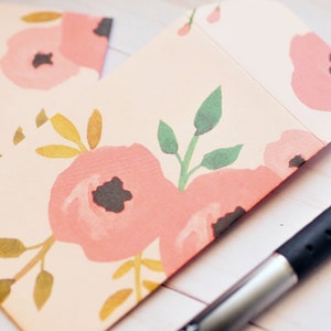 May include: A pink and white floral patterned envelope with a silver mechanical pencil on a white wooden surface.