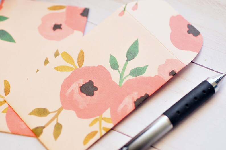 May include: A pink and white floral patterned envelope with a silver mechanical pencil on a white wooden surface.