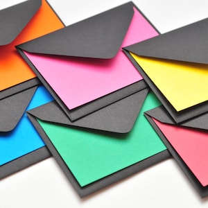 May include: Six colorful envelopes with black paper backing. The envelopes are layered on top of each other, with the top envelope being orange, followed by pink, yellow, blue, green, and red.