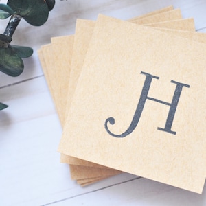 May include: A stack of square, light brown paper cards with the letter "H" printed in dark gray. The cards are arranged on a white wooden surface, with a sprig of green eucalyptus leaves in the background.