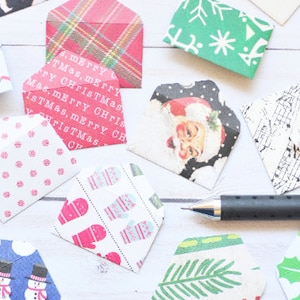 May include: A collection of small, decorative paper envelopes with various holiday-themed patterns. Designs include Santa Claus, mittens, snowflakes, and plaid. A pen rests near the envelopes, suggesting a crafting or stationery theme.