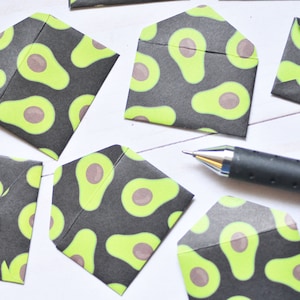 May include: A collection of small, black envelopes with a green avocado pattern. The envelopes are scattered on a white surface with a black pen.