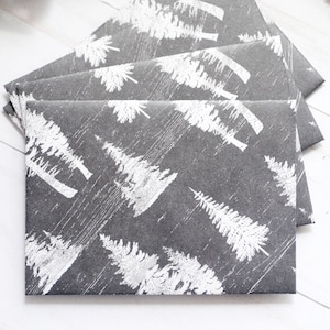 May include: A stack of gray envelopes with a white tree pattern. A black pen with silver accents rests on a white surface next to the envelopes. Greenery is visible in the background.