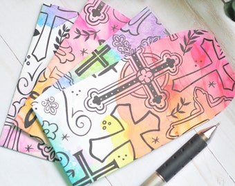 May include: Three colorful envelopes with a watercolor background and black cross designs. The envelopes are stacked on top of each other, with the top envelope being pink, the middle envelope being green, and the bottom envelope being blue. A black pen is lying on top of the envelopes.
