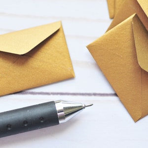 May include: A close-up of a black pen lying on a white surface with several gold envelopes scattered around it.