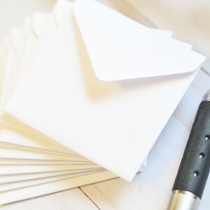 May include: A stack of white envelopes with a silver pen on a white wooden surface.