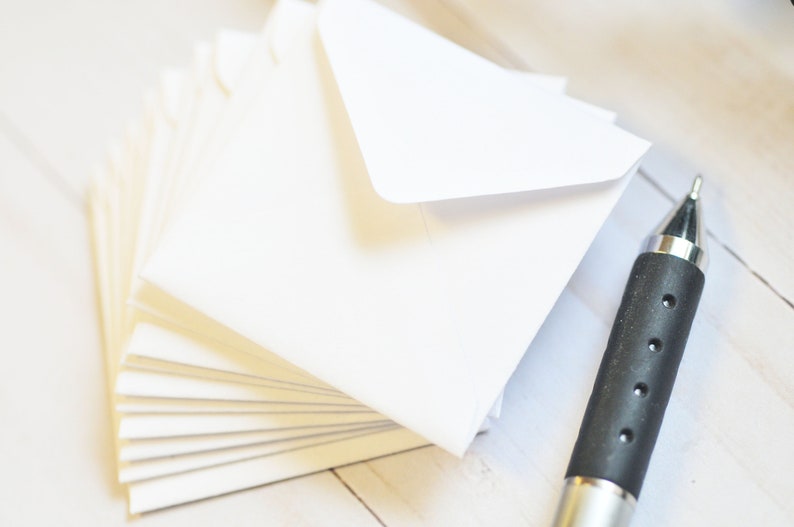 May include: A stack of white envelopes with a silver pen on a white wooden surface.