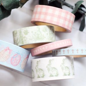 Peut inclure: Plusieurs rouleaux de ruban adhésif décoratif washi aux couleurs pastel. Les motifs comprennent des œufs de Pâques, des lapins, des croix et des carreaux vichy. Un rouleau porte l'inscription "easter blessings".