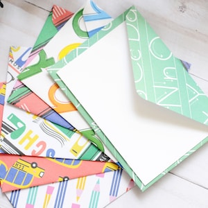 May include: A stack of colorful envelopes with school-themed designs, including a school bus, pencils, and books. The envelopes are folded and ready to be used for sending letters or notes.