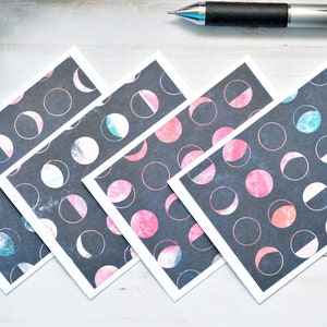 May include: Four square cards with a dark gray background and a pattern of white circles containing pink, blue, and white crescent moon phases. A silver pen and a sprig of greenery are in the background.