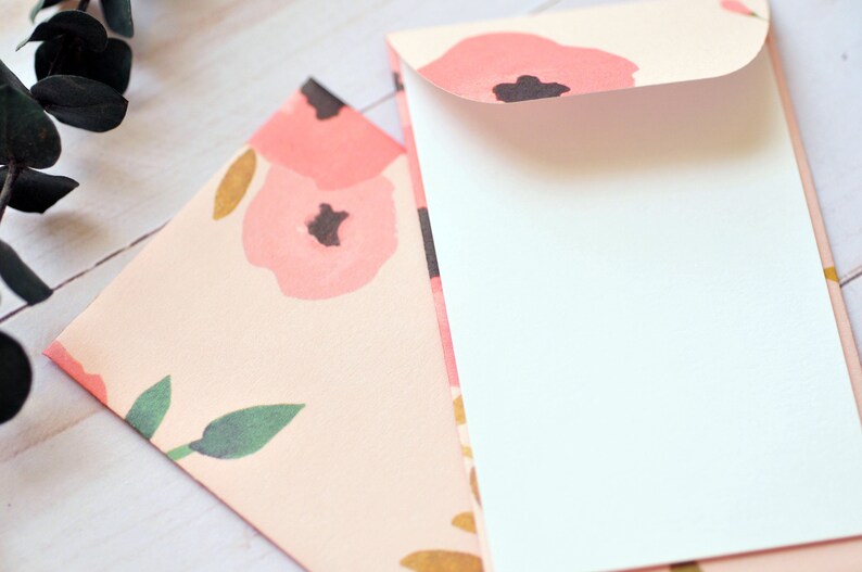 May include: Two envelopes, one with a floral pattern and the other with a white interior. The floral envelope has a pink background with pink and gold flowers and green leaves.