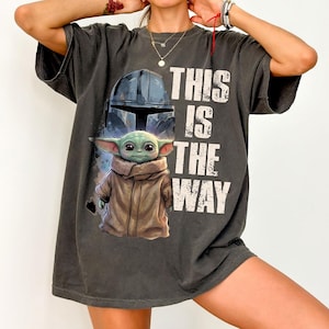 May include: Dark grey t-shirt with a graphic of The Child (Grogu) from The Mandalorian, the text "THIS IS THE WAY", and a helmet design. The shirt has short sleeves and a relaxed fit.
