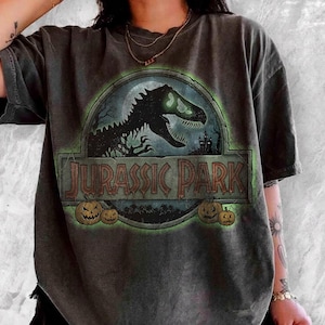 May include: A charcoal grey short-sleeve t-shirt featuring a circular graphic of a black dinosaur silhouette with green glowing eyes. The text JURASSIC PARK is printed in a weathered font, surrounded by small orange jack-o'-lantern pumpkins.