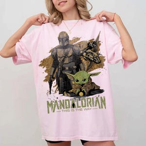 May include: A light pink t-shirt featuring a graphic of The Mandalorian and Grogu from the Star Wars series. The design includes the text "MANDALORIAN - THIS IS THE WAY" in green and white.
