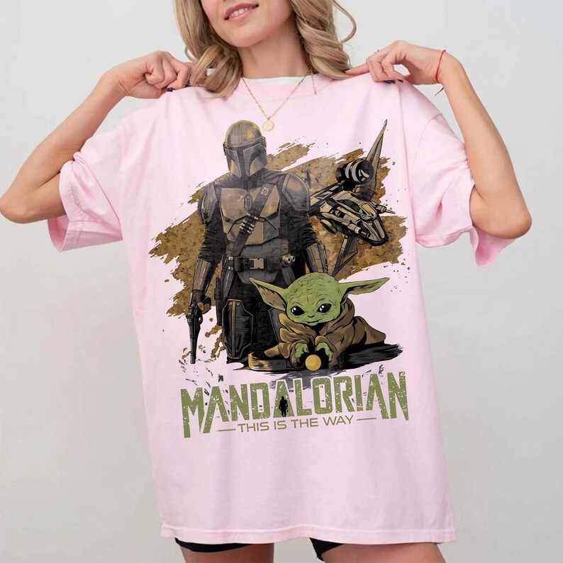 May include: A light pink t-shirt featuring a graphic of The Mandalorian and Grogu from the Star Wars series. The design includes the text "MANDALORIAN - THIS IS THE WAY" in green and white.