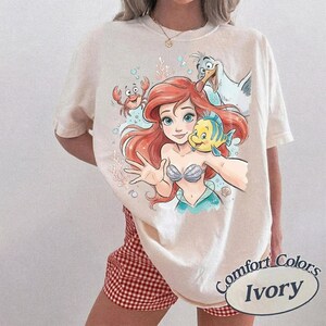 Op de afbeelding: Een ivoorkleurig t-shirt met korte mouwen en een grafische print van een zeemeermin met rood haar, een gele en blauwe vis, een rode krab en een witte meeuw. Het ontwerp bevat lichtblauwe bubbels en koraalaccenten op de voorzijde.
