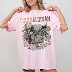 May include: Pink t-shirt featuring the word "MANDALORIAN" in gold lettering. The graphic includes the Mandalorian, Grogu, and a spaceship, surrounded by flowers. The shirt is a light pink color.