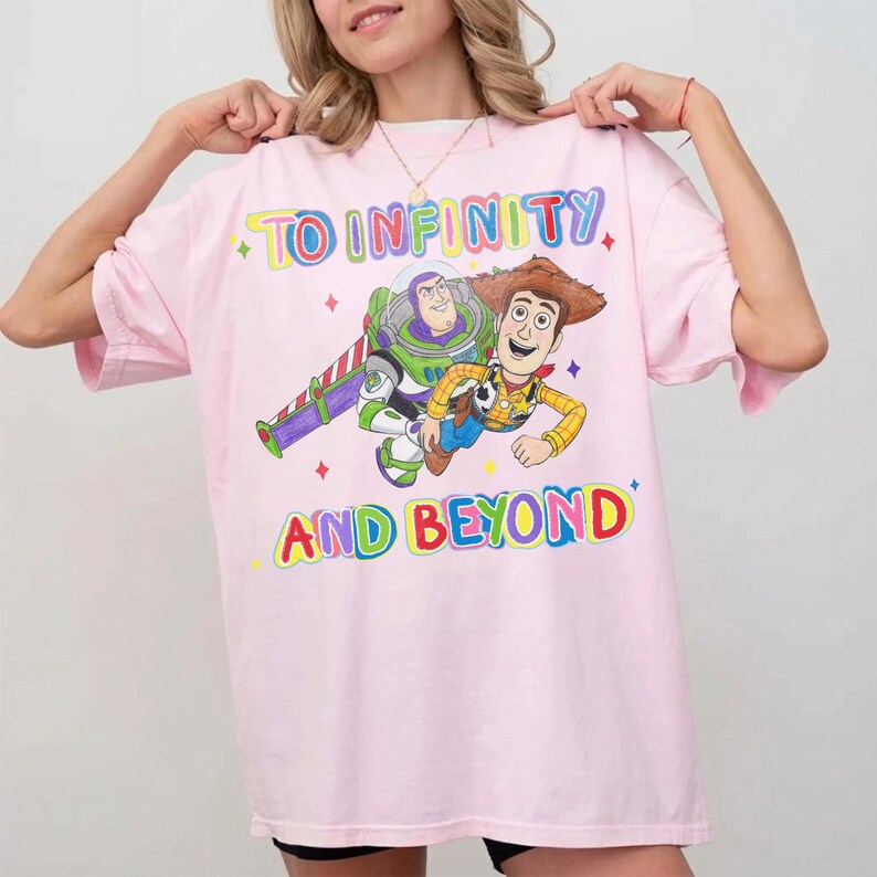 May include: A light pink t-shirt featuring a colorful graphic of Buzz Lightyear and Woody from Toy Story. The text "TO INFINITY" and "AND BEYOND" are printed above and below the characters in a rainbow font. The shirt has short sleeves.