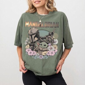 May include: Olive green t-shirt featuring a graphic of The Mandalorian, Grogu, and a spaceship, surrounded by flowers. The word "MANDALORIAN" is printed across the top in a gradient font. The shirt has short sleeves and a relaxed fit.