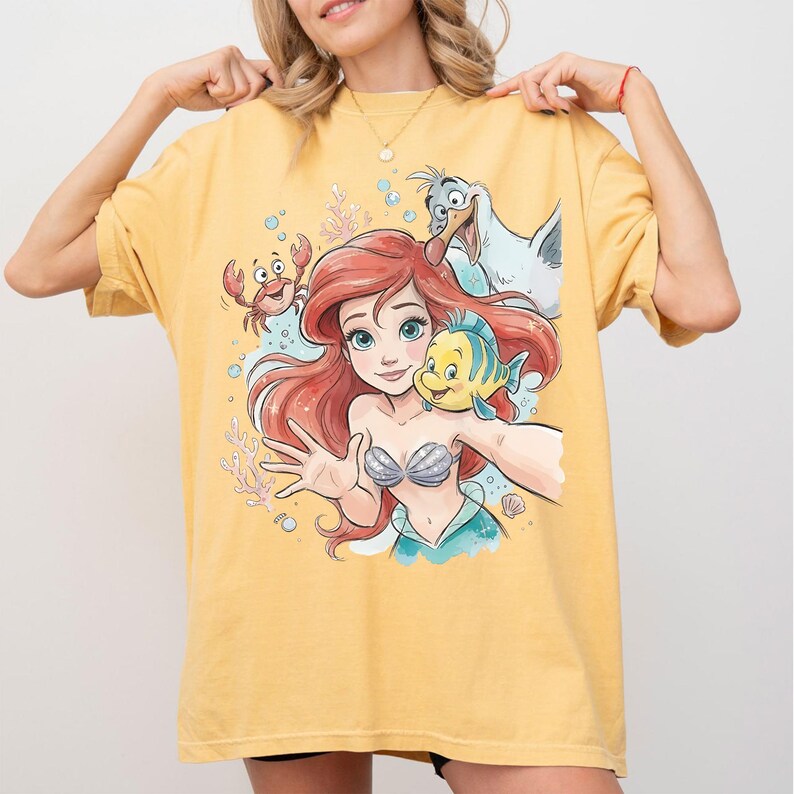 Op de afbeelding: Een mosterdgeel T-shirt met korte mouwen en een grafische print van een zeemeermin met rood haar, een kleine gele en blauwe vis, een rode krab en een witte meeuw. Het ontwerp bevat onderwaterelementen zoals bubbels en koraal.