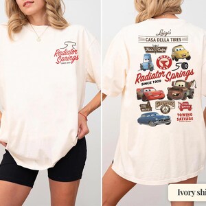 May include: An ivory t-shirt featuring a small Radiator Springs logo on the front left chest. The back displays a large graphic with various Cars characters, including Lightning McQueen and Mater, alongside text reading Luigi's Casa Della Tires.