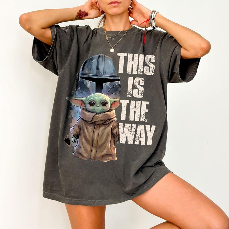 May include: Dark grey t-shirt with a graphic of The Child (Grogu) from The Mandalorian, the text "THIS IS THE WAY", and a helmet design. The shirt has short sleeves and a relaxed fit.