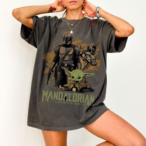 May include: Dark gray t-shirt featuring a graphic of The Mandalorian, Grogu, and a spaceship. The text "MANDALORIAN - THIS IS THE WAY" is printed below the image. The shirt has short sleeves and a relaxed fit.