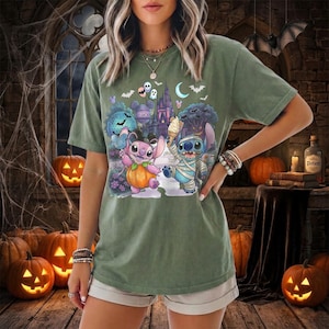 May include: An olive green short-sleeve t-shirt featuring a graphic print of two cartoon characters, one dressed as a pumpkin and the other as a mummy, standing in front of a castle with bats, skulls, and a crescent moon in the background.