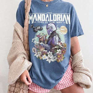 May include: Blue jean-coloured t-shirt featuring a graphic of The Mandalorian and Grogu surrounded by flowers, with the text "THE MANDALORIAN THIS IS THE WAY." The shirt also has the text "COMFORT COLORS BLUE JEAN."