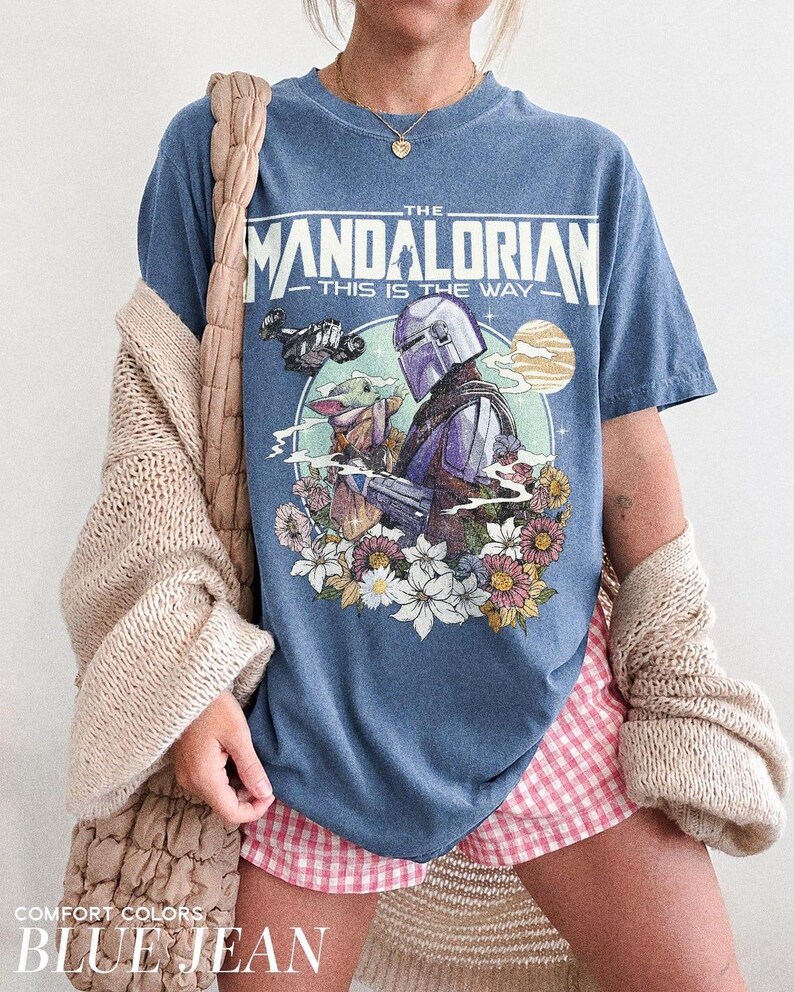 May include: Blue jean-coloured t-shirt featuring a graphic of The Mandalorian and Grogu surrounded by flowers, with the text "THE MANDALORIAN THIS IS THE WAY." The shirt also has the text "COMFORT COLORS BLUE JEAN."