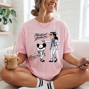 K&ouml;nnte beinhalten: Hellrosa T-Shirt mit einer Cartoon-Illustration von Michael Jackson und Mickey Mouse. Die Texte "Michael Jackson" und "Mickey Mouse" sind in schwarzer Schrift aufgedruckt. Das T-Shirt ist ein l&auml;ssiger, &uuml;bergro&szlig;er Stil.