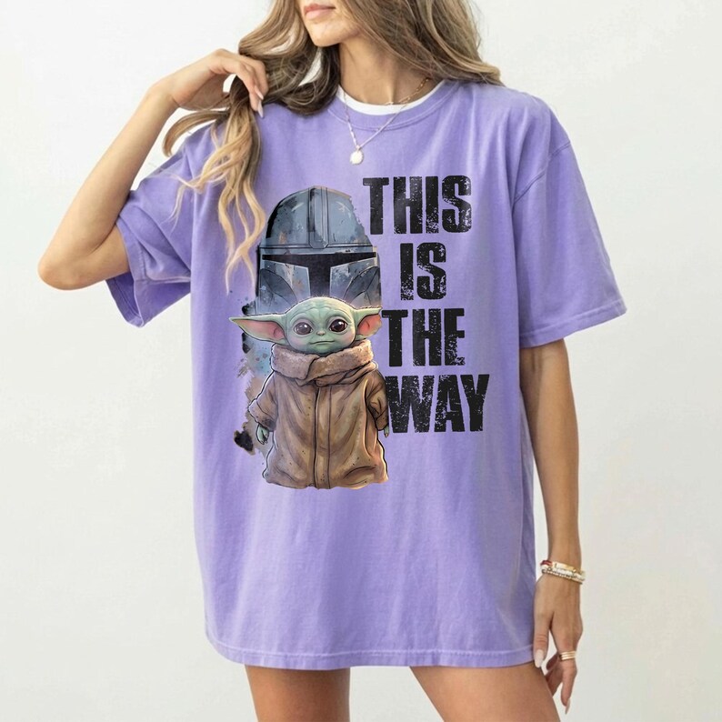 May include: A lilac t-shirt with a graphic of a helmet and a character with large ears wearing a brown coat. The text "THIS IS THE WAY" is printed in black on the front of the shirt.
