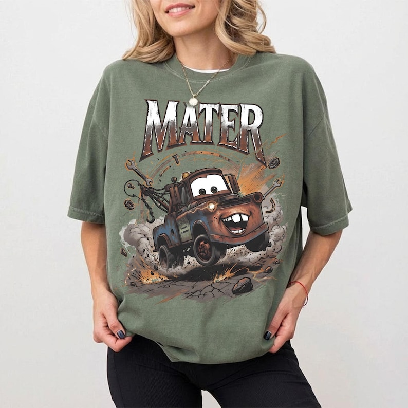 May include: Olive green t-shirt featuring a cartoon image of Mater from the movie Cars. The word "MATER" is printed above the image in a distressed, rusty font. The graphic shows Mater with a big smile, kicking up dust and debris.