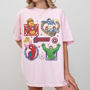 Puede incluir: Una camiseta rosa claro con personajes de dibujos animados disfrazados de superh&eacute;roes. El dise&ntilde;o incluye a Winnie the Pooh como Iron Man, un burro como Thor, Spider-Pig como Spider-Man y Hulk. La palabra "Avengers" est&aacute; impresa en rojo.