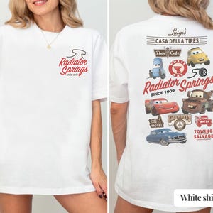 May include: A white t-shirt featuring a small Radiator Springs logo on the front left chest. The back displays a large graphic with various Cars characters, including Lightning McQueen and Mater, alongside text reading Luigi's Casa Della Tires.