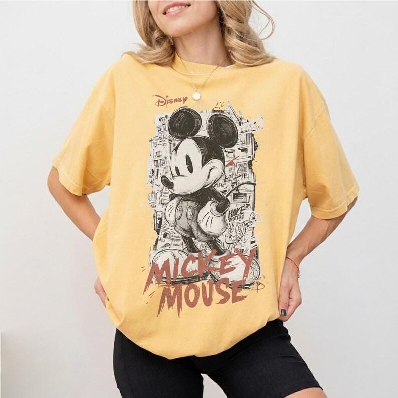 Puede incluir: Camiseta amarilla mostaza con un gr&aacute;fico en blanco y negro de Mickey Mouse. El dise&ntilde;o incluye el texto "Mickey Mouse" en una fuente desgastada. El logotipo de Disney es visible. La camiseta tiene un corte holgado y extragrande.