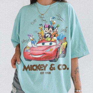 May include: A light blue t-shirt featuring a graphic of Disney characters, including Mickey Mouse, Minnie Mouse, and Donald Duck, on a red race car. The shirt has the text "Mickey & Co. EST 1928".